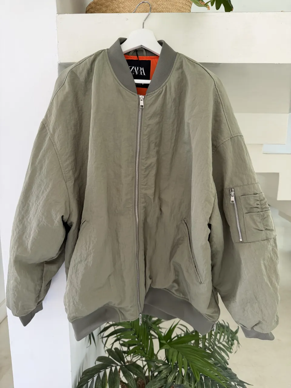 Campera bomber color verde militar con forro naranja. Tiene cierre al frente, bolsillos laterales y un bolsillo con cierre en la manga izquierda.
