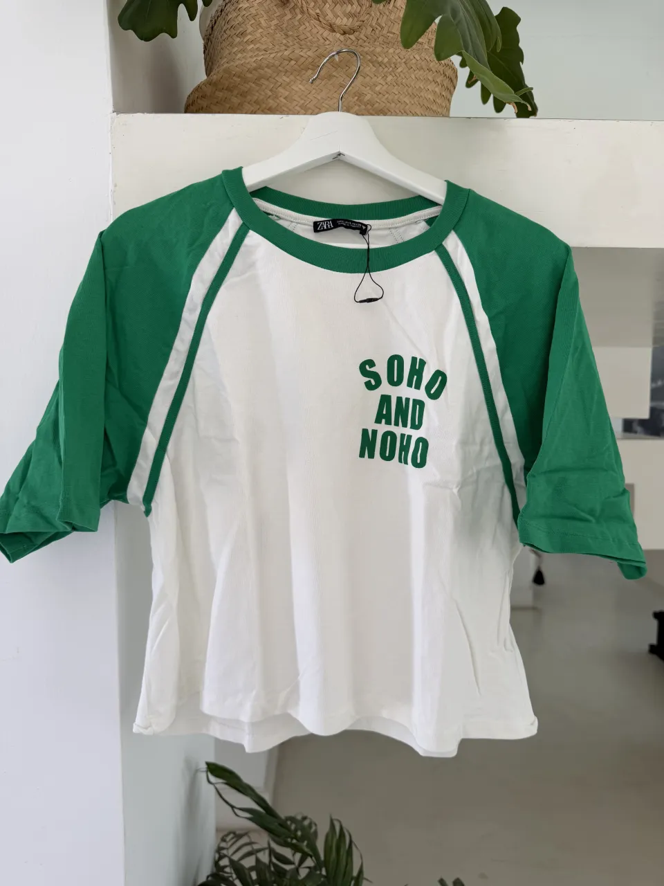 Remera blanca con mangas cortas de color verde. Tiene un cuello redondo y un estampado en el frente con la inscripción "SOHO AND NOHO" en color verde.
