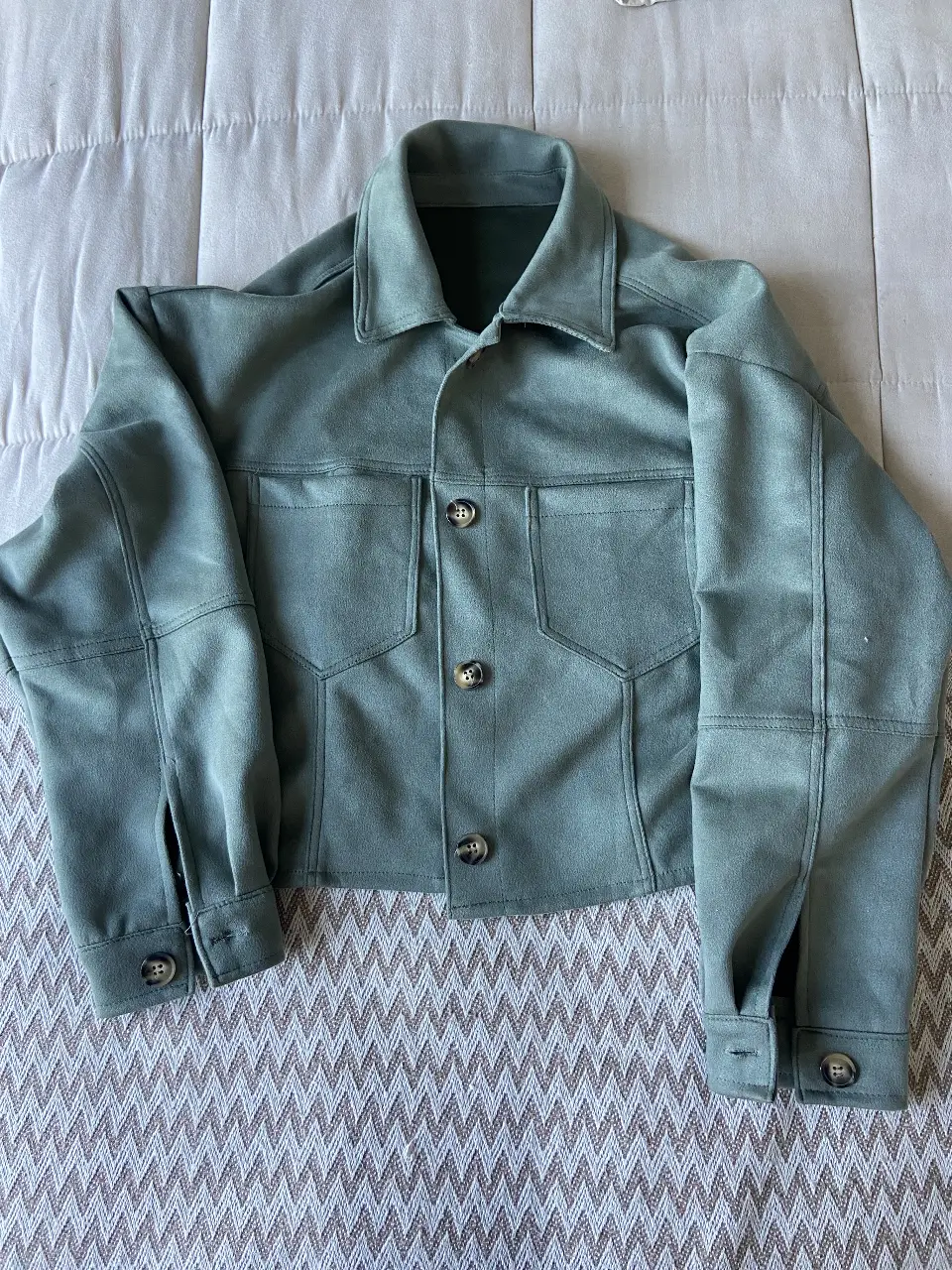 Campera estilo bomber de gamuza sintética en color verde. Posee dos bolsillos delanteros con solapa y botones, y botones en los puños. Ideal para un look casual y moderno.