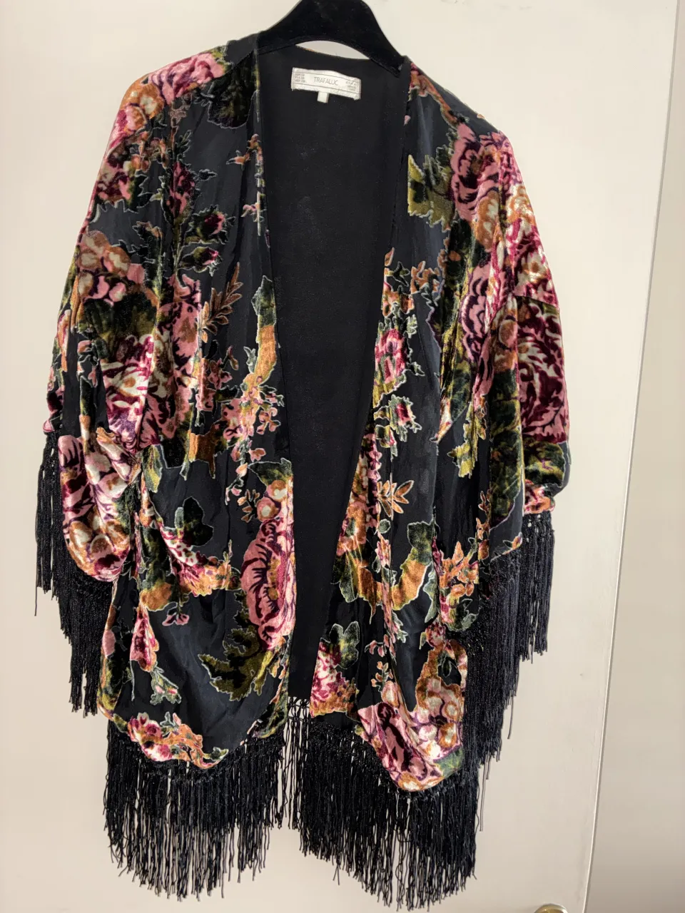 Kimono negro con estampado floral en tonos rosas, verdes y ocres. Posee flecos largos en mangas y ruedo.