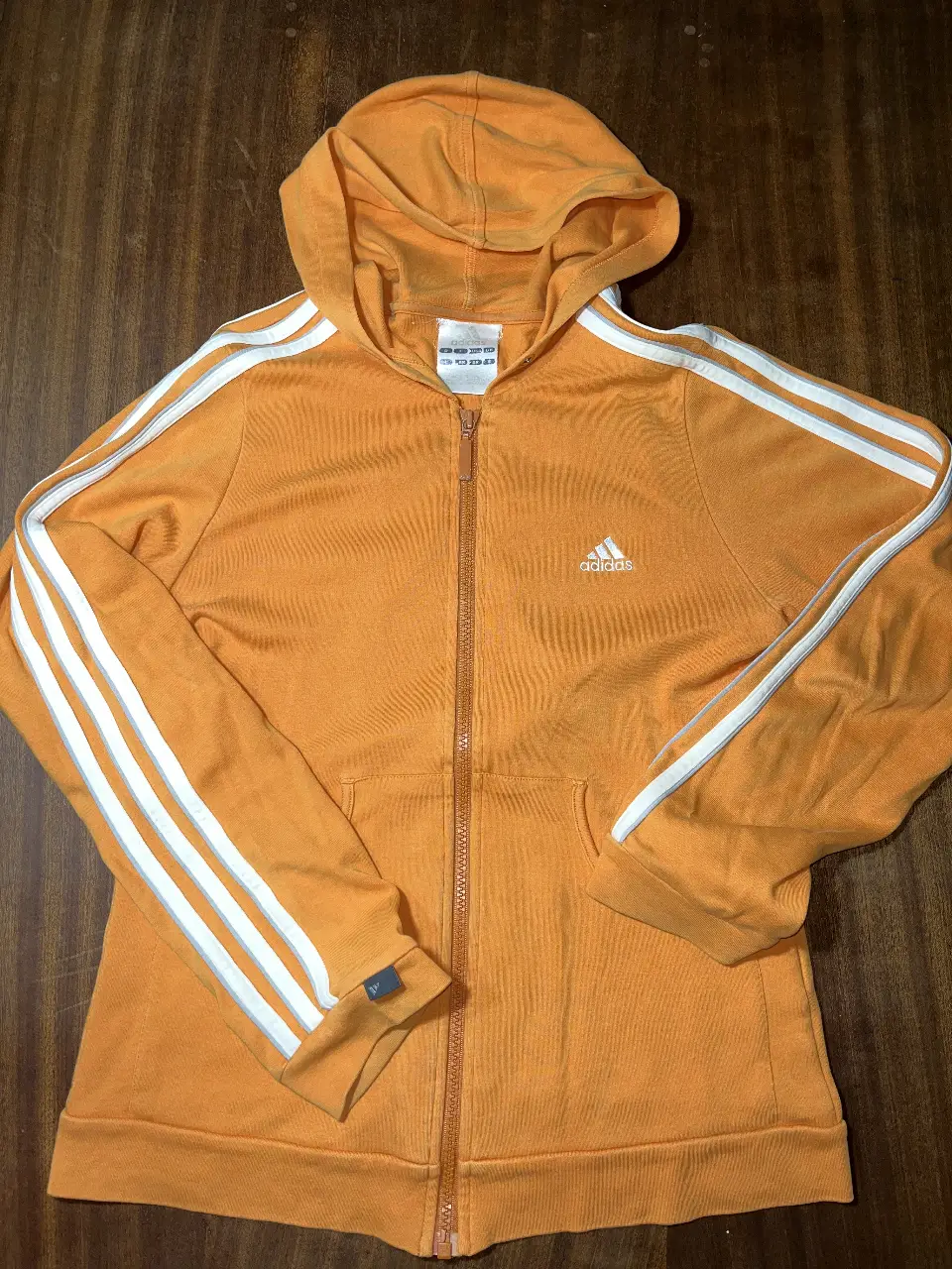 Buzo con cierre Adidas naranja con las tres tiras blancas en las mangas. Ideal para un look deportivo y casual.
