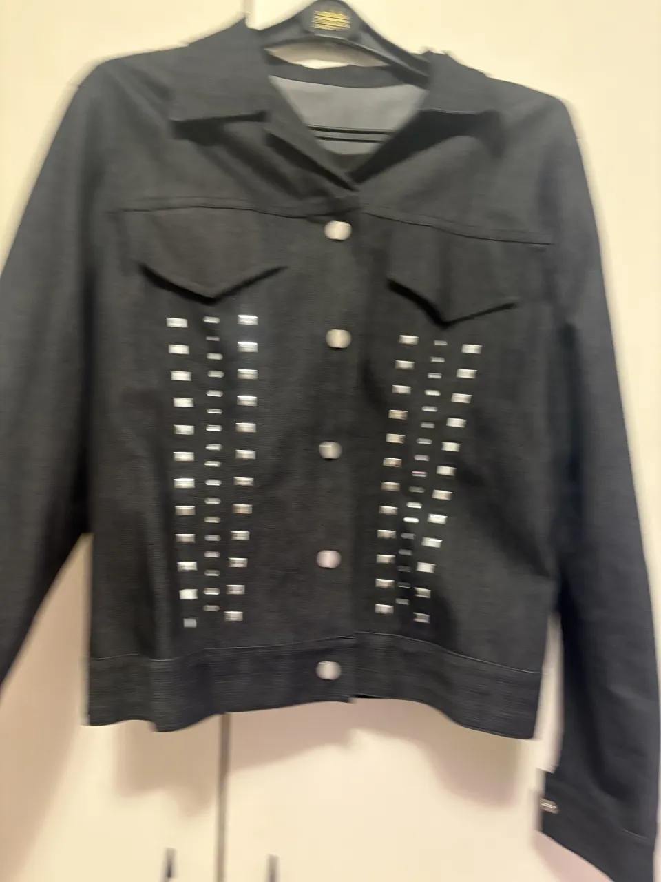 Campera de jean negra con tachas plateadas cuadradas dispuestas en dos filas verticales en el frente. Posee cuello camisero, botones al frente y bolsillos con solapa en el pecho. tengo el pantalón haciendo juego 