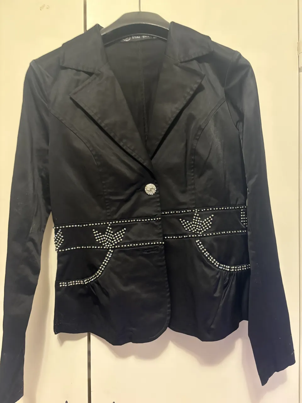 Blazer negro con detalles de strass en los bolsillos delanteros y en la cintura. La parte trasera tiene un lazo de strass.