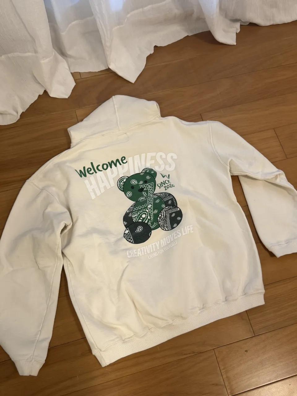Buzo blanco con capucha y estampa de oso de peluche con pañuelo estilo bandana en verde y negro. La estampa dice "Welcome HAPPINESS" y "CREATIVITY MOVES LIFE LIVING FOR YOURSELF".