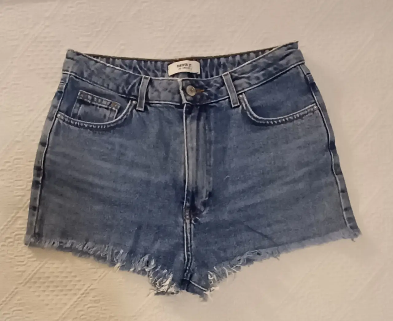 Short de jean azul de mujer, tiro alto, con detalles desflecados en el borde inferior. Ideal para un look casual y moderno.