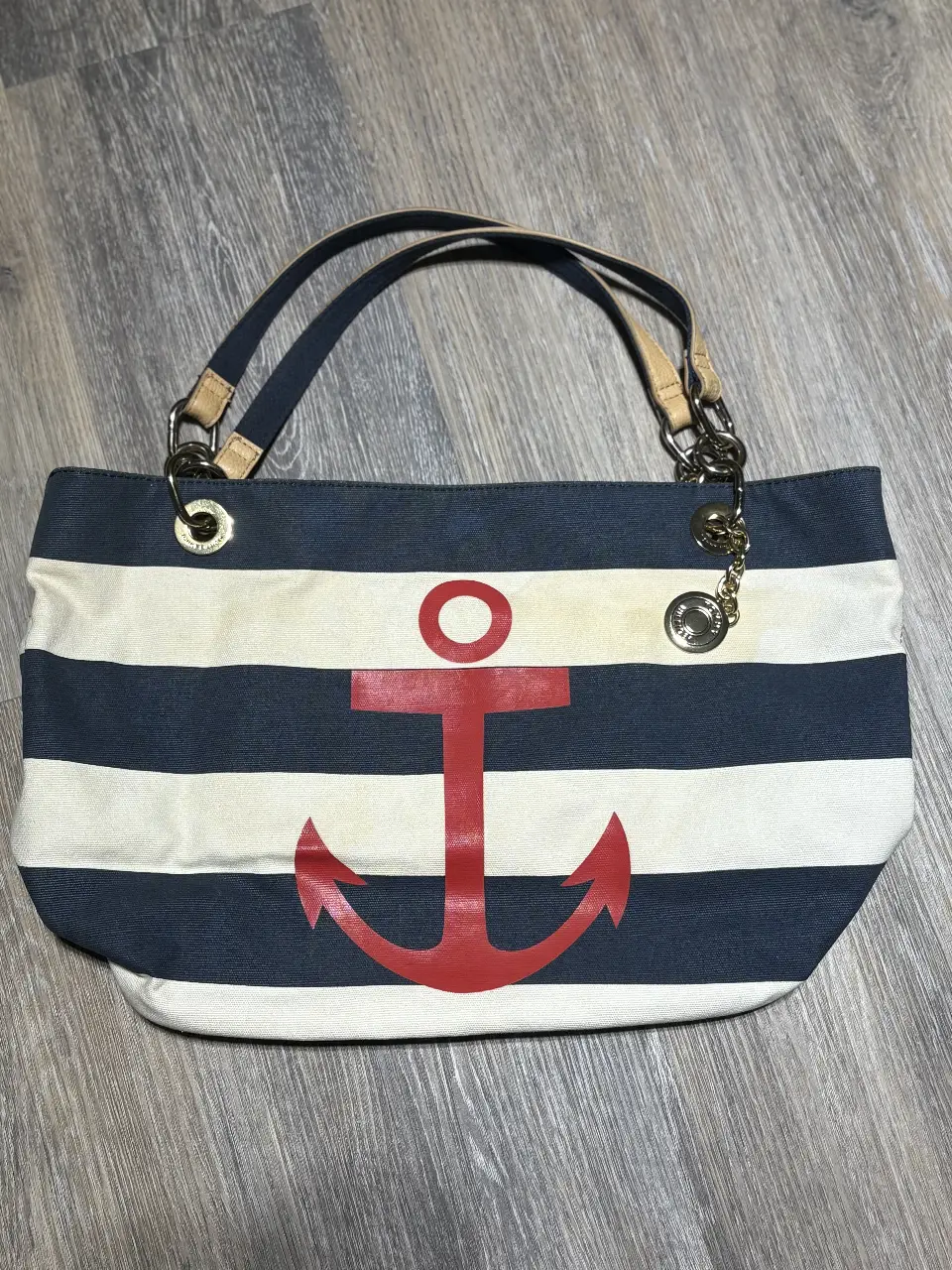 Bolso tote de lona a rayas azul y blanco con diseño de ancla roja. Cuenta con manijas de cuero y detalles dorados. Interior espacioso con bolsillo interno y cierre. Ideal para playa o uso diario.