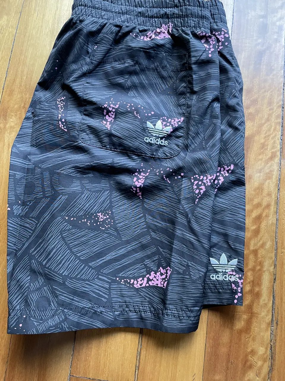 Short deportivo Adidas para hombre, con estampado geométrico en tonos grises y detalles en rosa. Ideal para playa, pile y uso casual.
