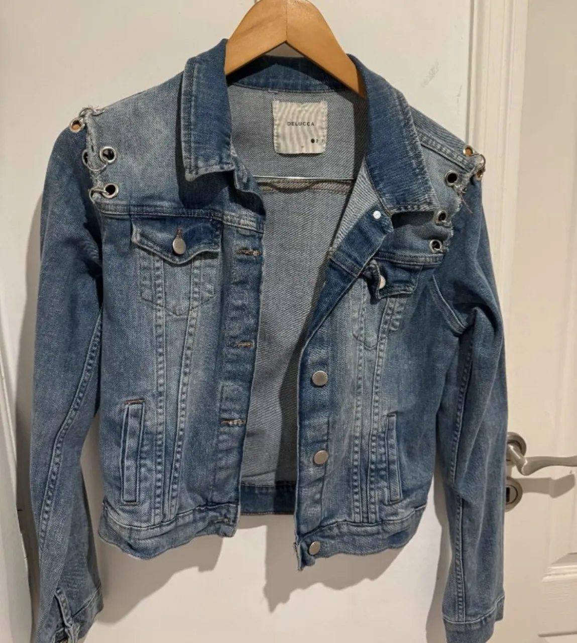 Campera de jean azul con detalles de ojales y argollas metálicas en los hombros. Posee bolsillos frontales con botones.