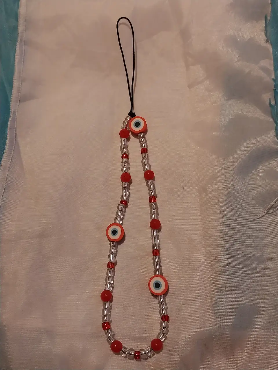 Llavero artesanal con cuentas rojas y blancas, adornado con ojos turcos y un cordón negro. Perfecto para agregar un toque de estilo y protección a tu celu
