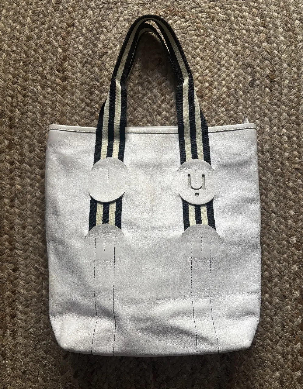 Bolso tote de cuero blanco con manijas a rayas negras y blancas. Detalles de diseño con apliques circulares y letras "U". Ideal para un look casual y moderno.