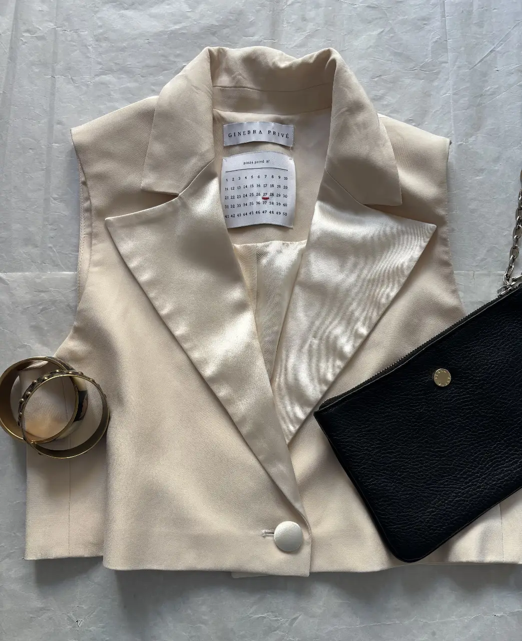 Chaleco sastrero color crudo de la marca Ginebra Privé. Confeccionado con un tejido satinado, este chaleco sin mangas presenta un diseño elegante con solapas y un solo botón. Ideal para un look sofisticado y moderno.