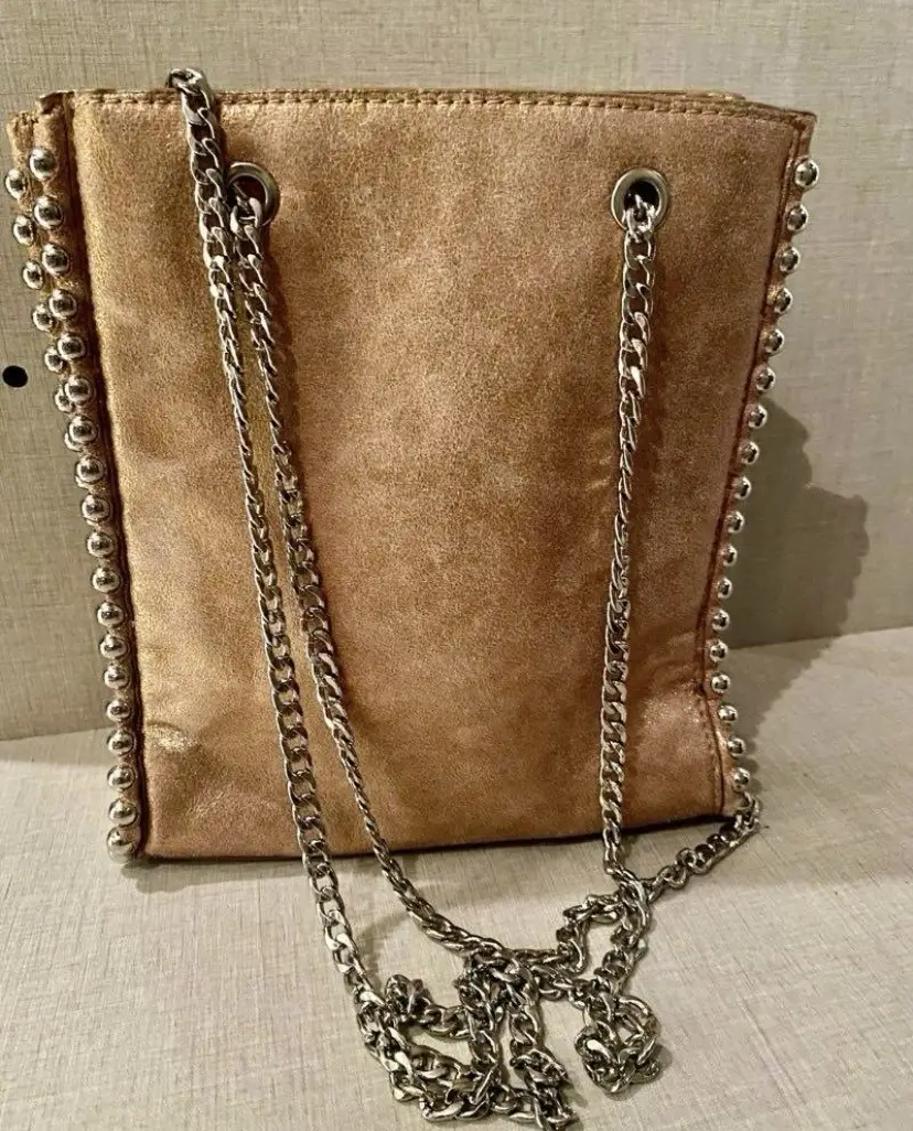 Cartera dorada con tachas plateadas en los bordes y cadena plateada. Ideal para darle un toque de glamour a cualquier look.