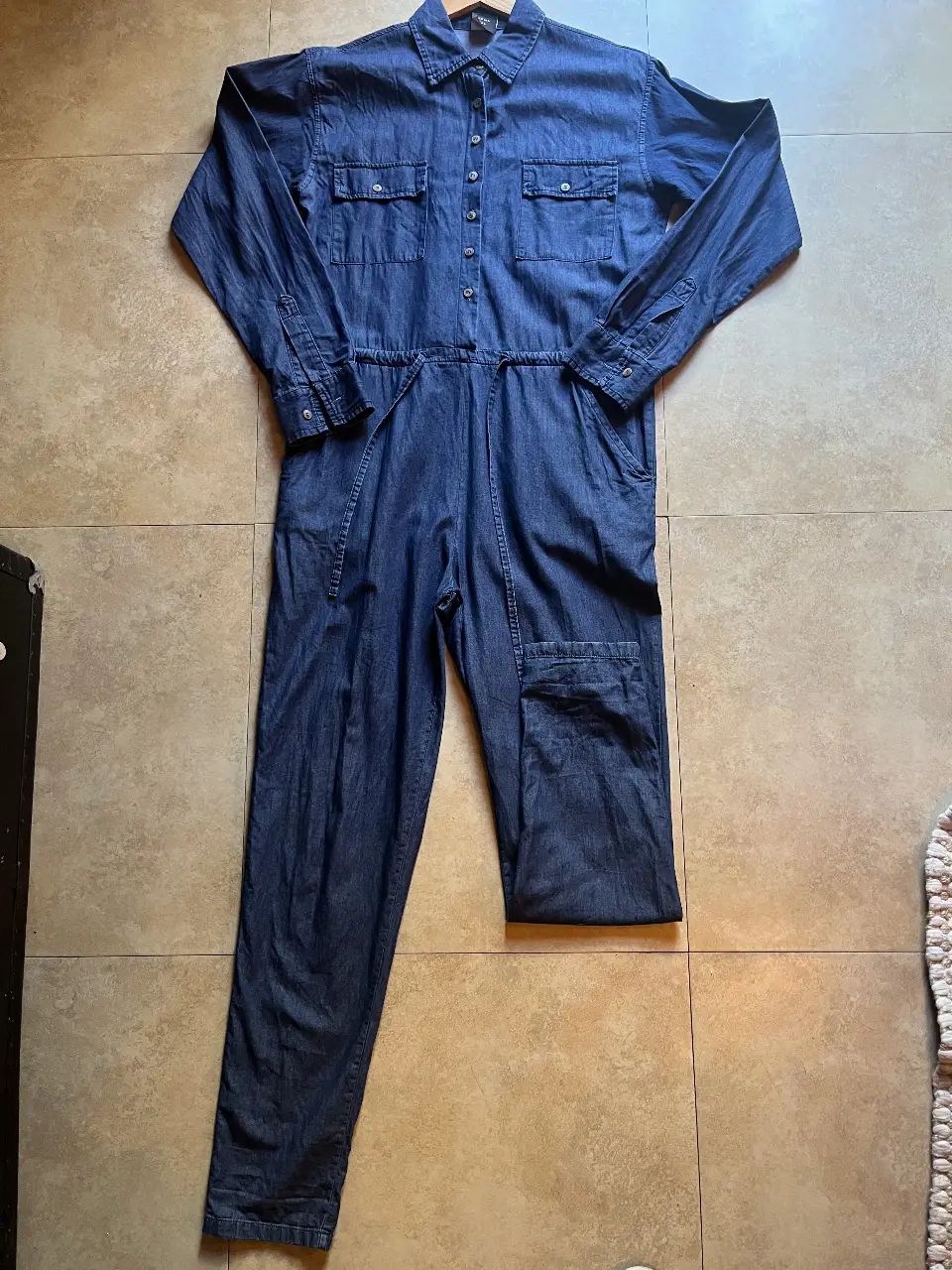 Mono largo de jean azul oscuro, con mangas largas y botones. Tiene bolsillos en el pecho y en los laterales de los pantalones. Cintura con ajuste y detalle de costuras.