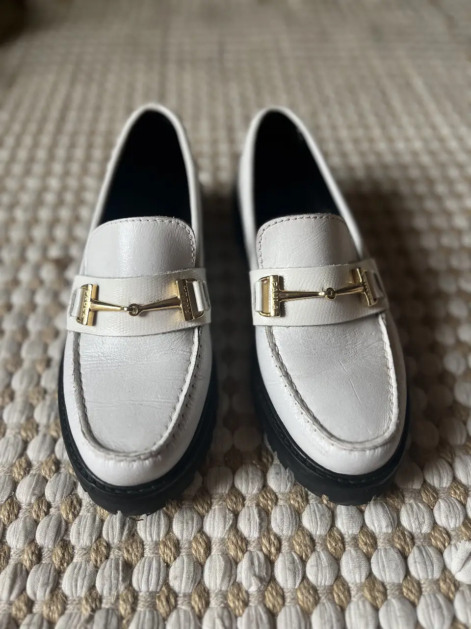 Mocasines blancos con detalle dorado, suela negra. Ideales para un look casual y elegante.