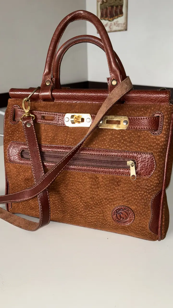 Cartera de mano de gamuza marrón con detalles en cuero. Posee correa para colgar y cierre dorado. Ideal para un look elegante y retro.
