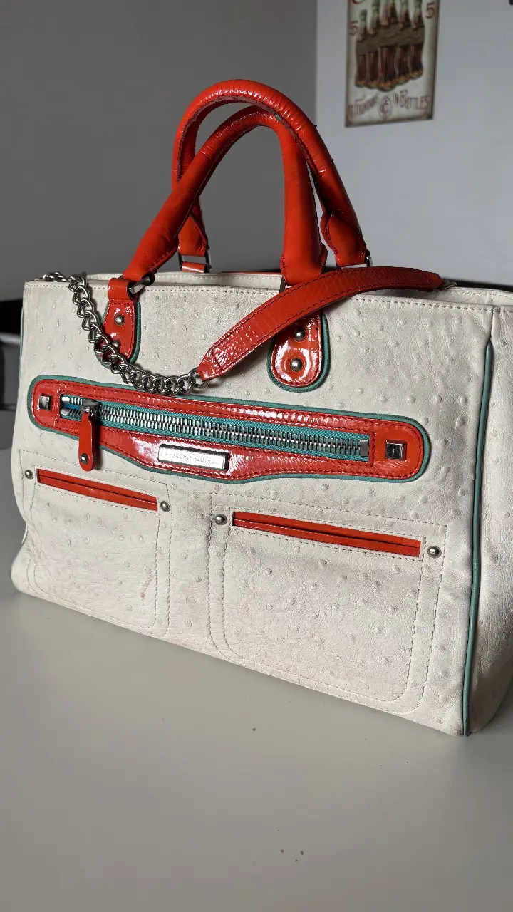 Cartera de mano blanca con detalles en naranja y turquesa. Diseño elegante con cadena plateada y manijas naranjas. Ideal para un look sofisticado.