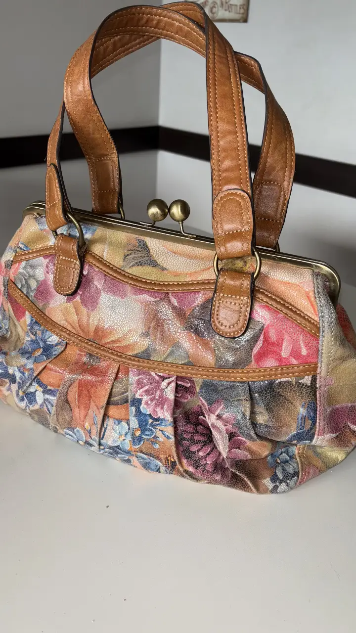 Cartera de mano con estampado floral vintage, detalles en cuero marrón y cierre metálico. Ideal para un look retro y femenino.