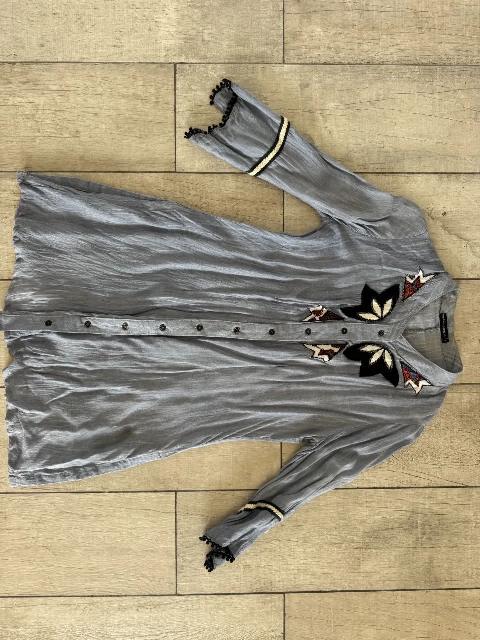 Camisa de manga larga color gris con detalles bordados en la pechera y puños. Los puños tienen un borde con pompones negros y una tira de detalles blancos y negros.