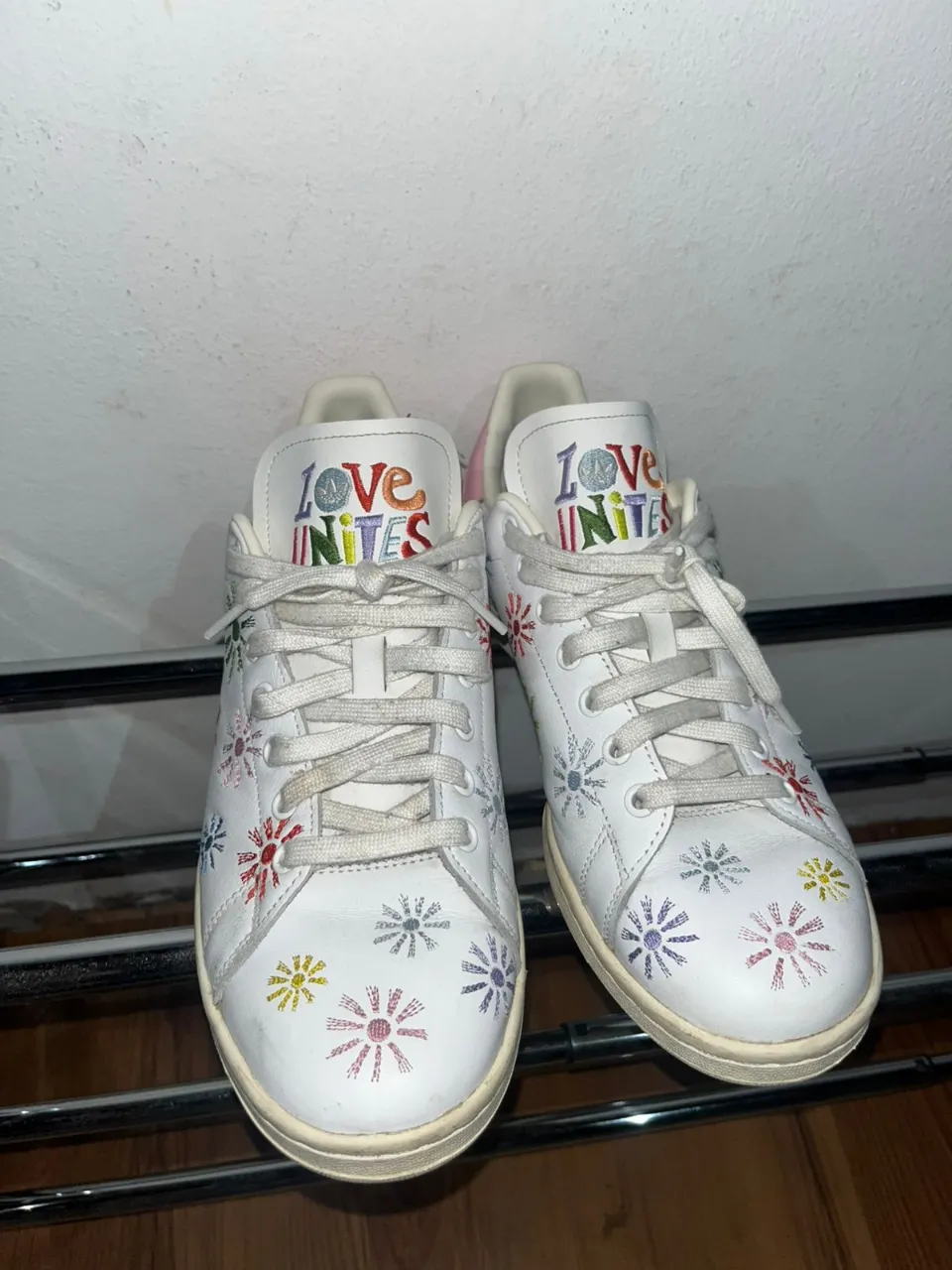 Zapatillas blancas con cordones y detalles de flores de colores bordadas en toda la superficie. En la lengüeta se lee "LOVE UNITES" con letras multicolores.