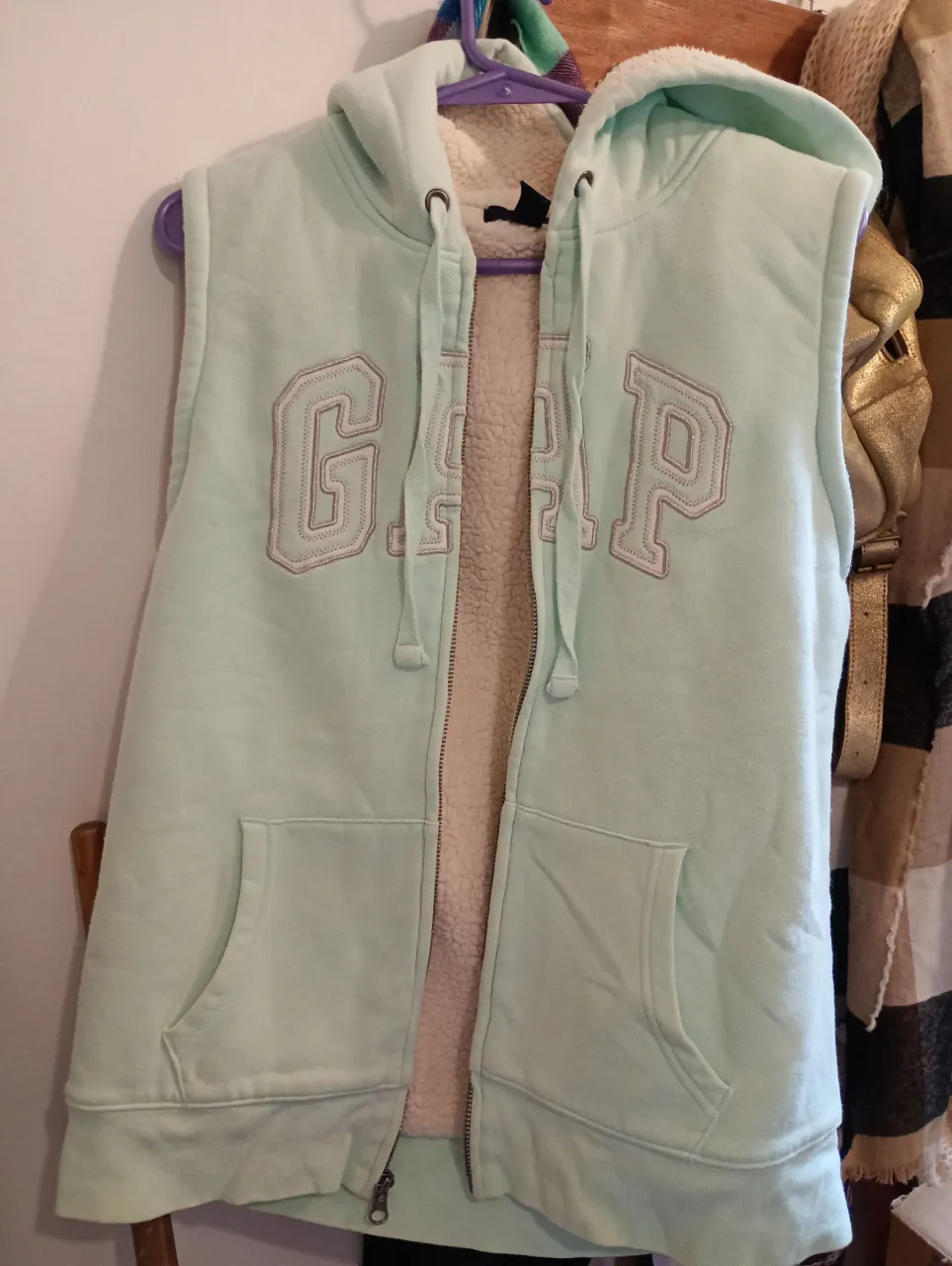 Buzo canguro sin mangas de color verde pastel con interior de sherpa blanco. Tiene capucha, bolsillos frontales y el logo de GAP bordado en el frente. Buen estado 