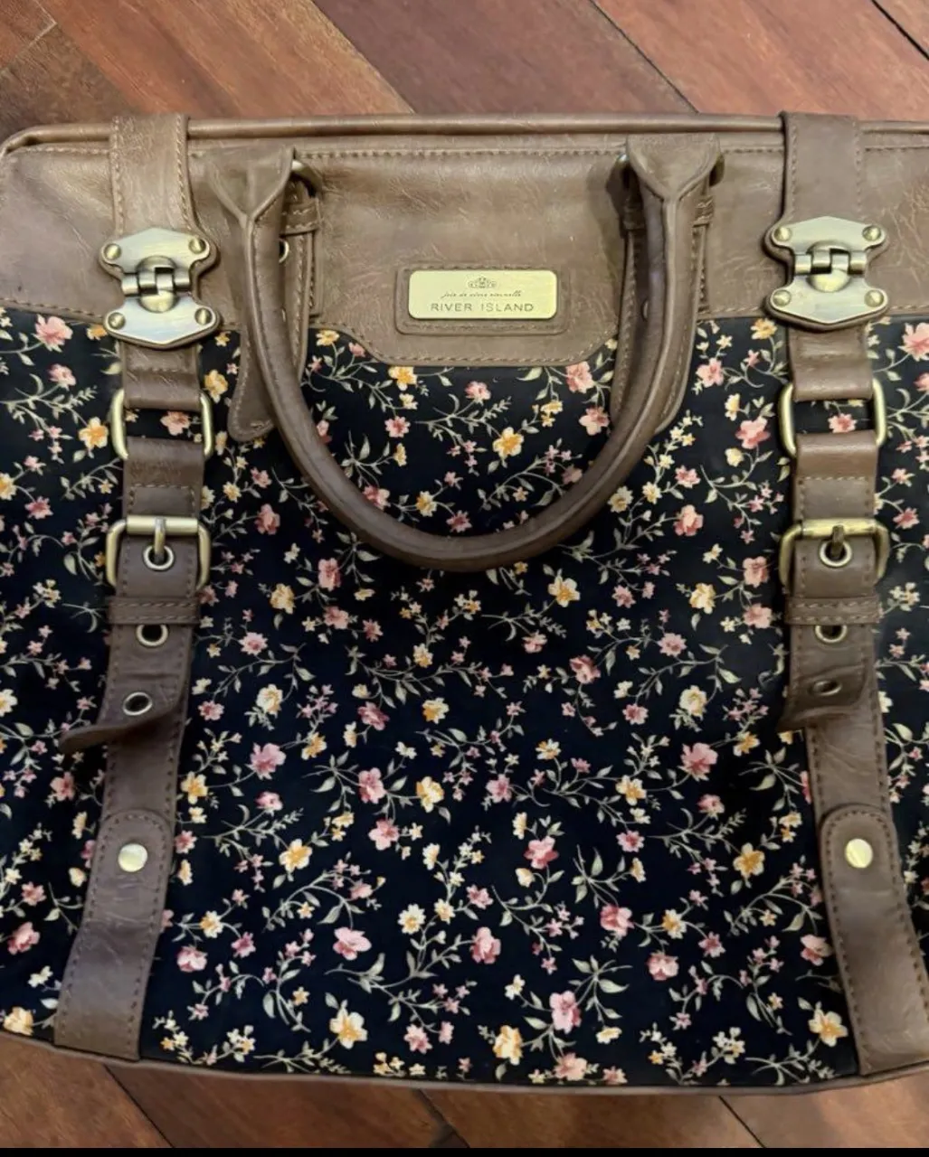 Bolso de mano con estampa floral sobre fondo negro. Posee detalles y manijas de cuero marrón y correa larga. Tiene herrajes metálicos dorados.