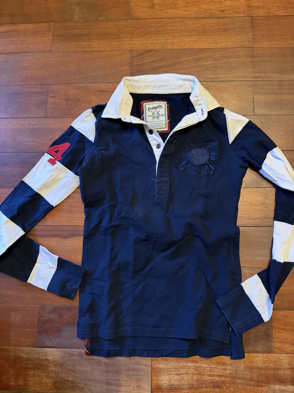 Remera manga larga, con cuello blanco y detalles a rayas en las mangas. Tiene un número 4 rojo en la manga izquierda y un bordado en el frente. Ideal para un look casual y deportivo. Trae botón de repuesto 