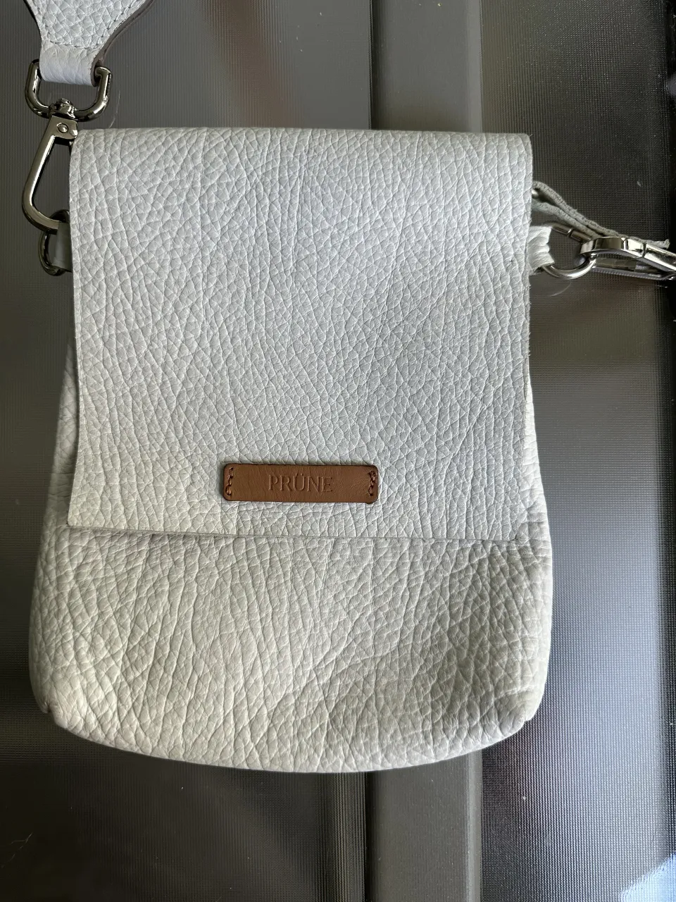 Cartera Prune de cuero blanca.Posee una correa de tela a rayas en tonos beige y blanco, y se puede usar cruzada o al hombro. Tiene cierre.