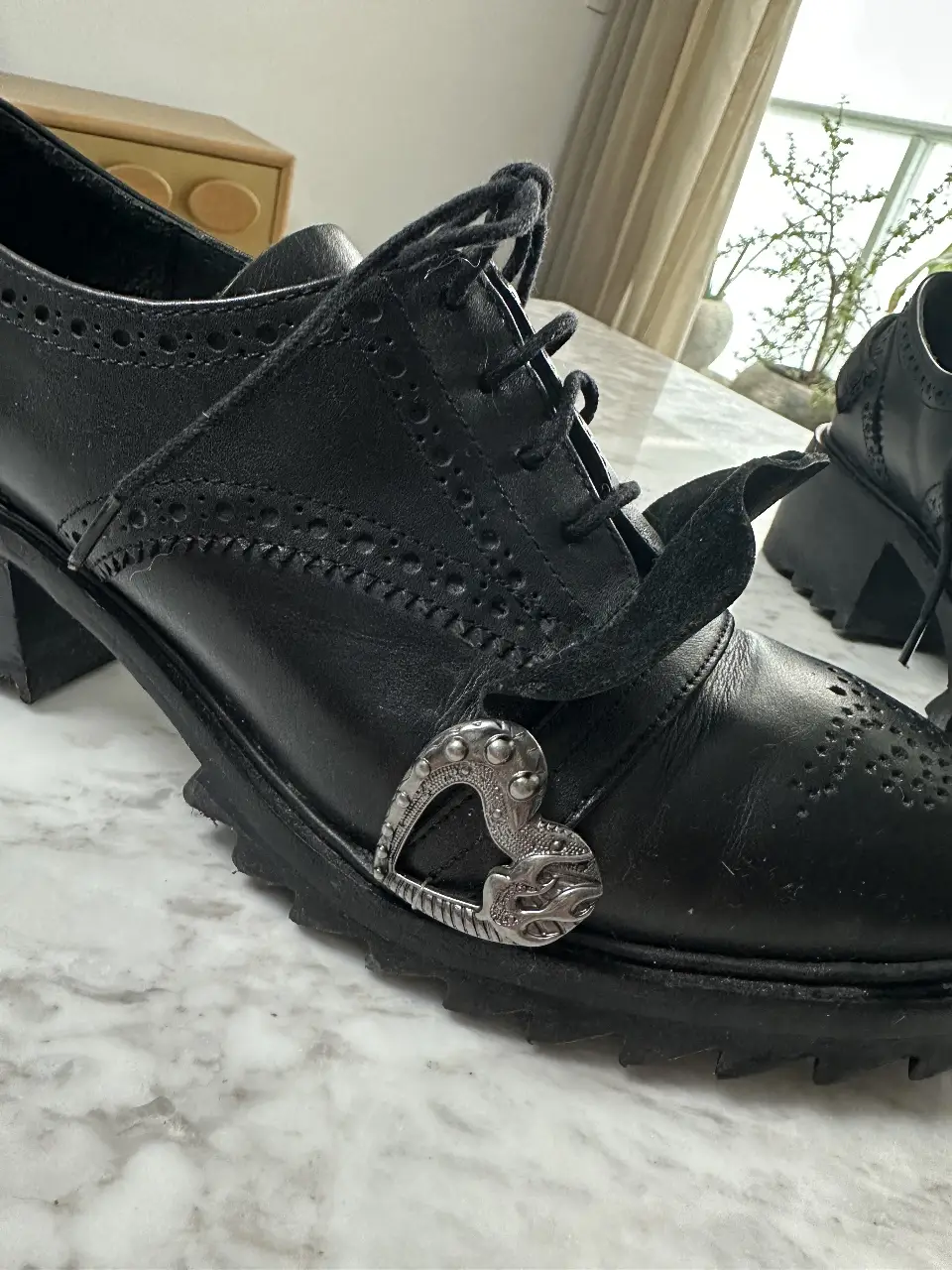 Botines negros de cuero con detalles de brogue y hebilla en forma de corazón. Tienen un diseño de plataforma y taco ancho.