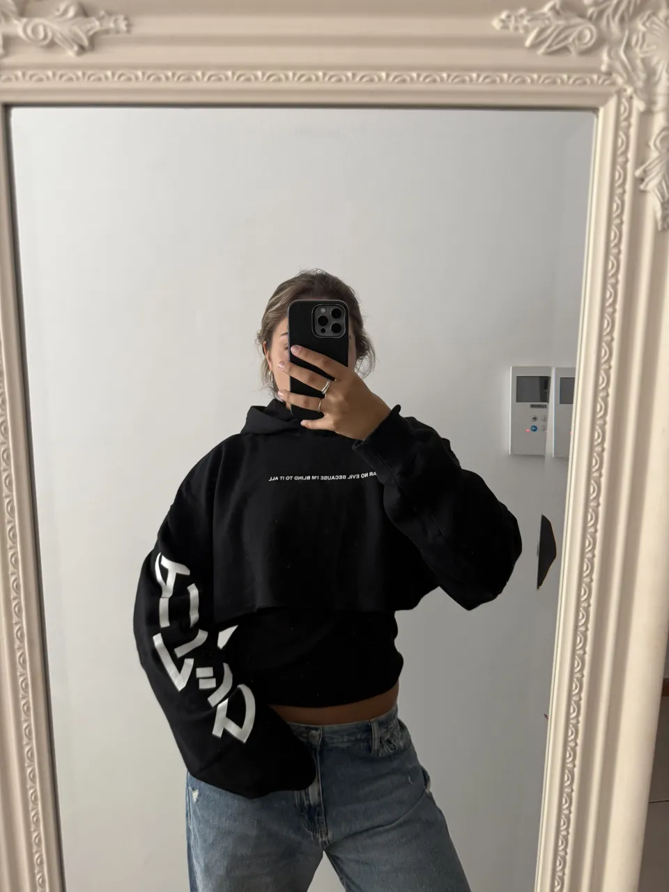 Buzo crop negro con capucha y mangas largas. Presenta estampa blanca en las mangas y en la parte delantera y trasera del cuello. La estampa en las mangas tiene un diseño de letras grandes y estilizadas. La estampa en la parte trasera del cuello es un texto más pequeño.