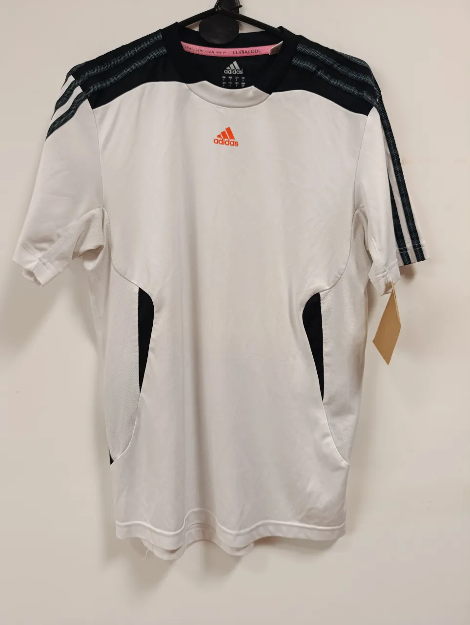 Remera deportiva blanca con detalles en negro y naranja. Ideal para entrenamientos o uso casual. Marca Adidas.