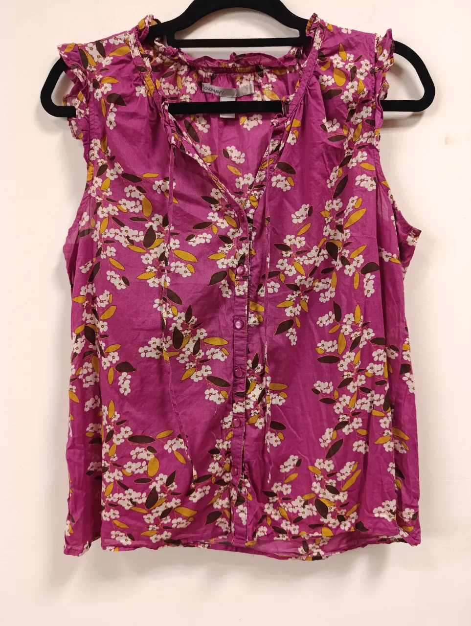 Blusa sin mangas, color fucsia vibrante con un delicado estampado floral en blanco, amarillo y marrón. Posee cuello V con cordones para atar, detalles de volados en los hombros y botones al frente. Ideal para un look fresco y femenino.