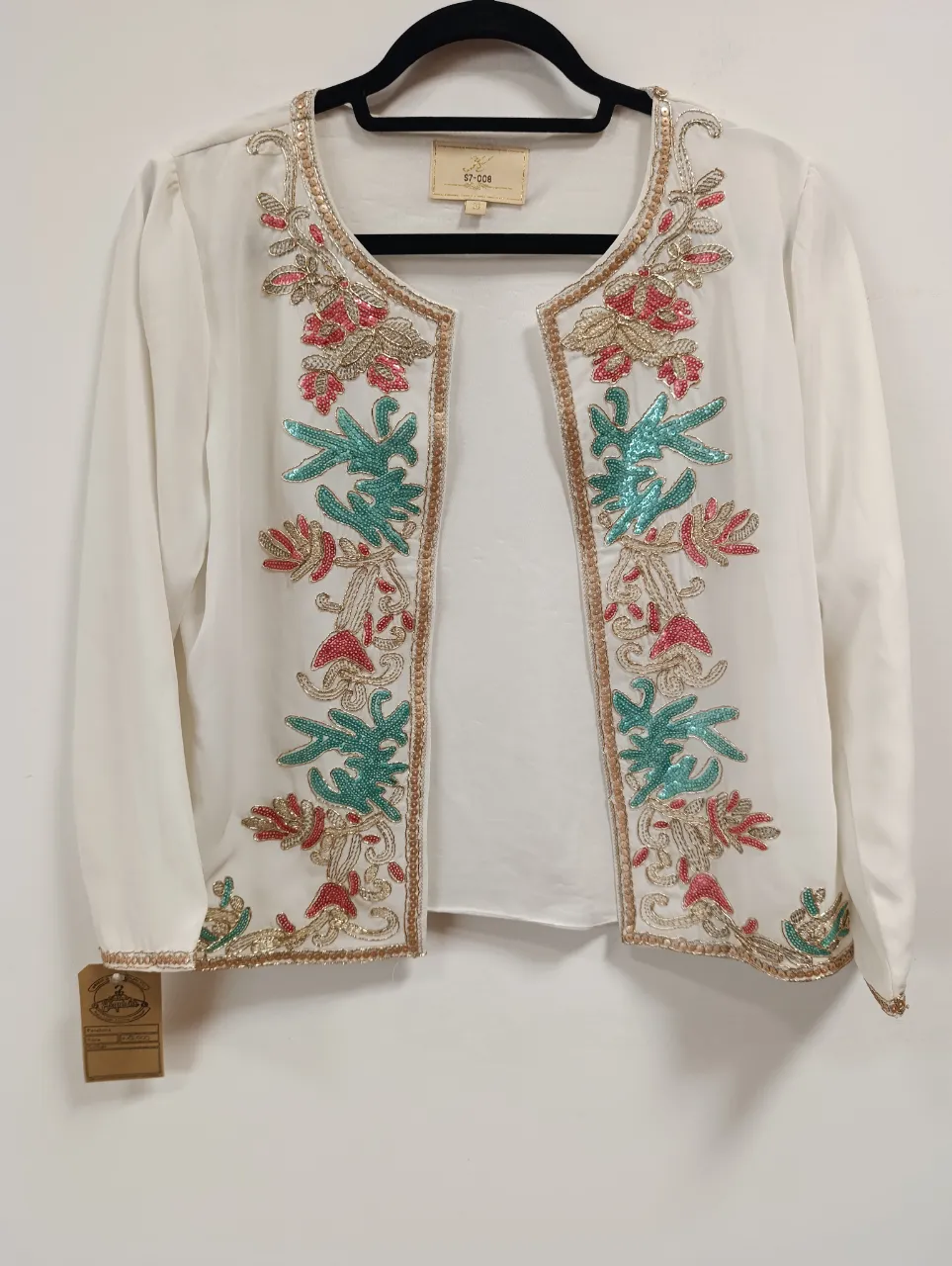 Blazer blanco con bordados florales y lentejuelas en tonos fucsia, verde y dorado. Posee mangas largas y un corte abierto al frente. Ideal para complementar un look elegante y sofisticado.