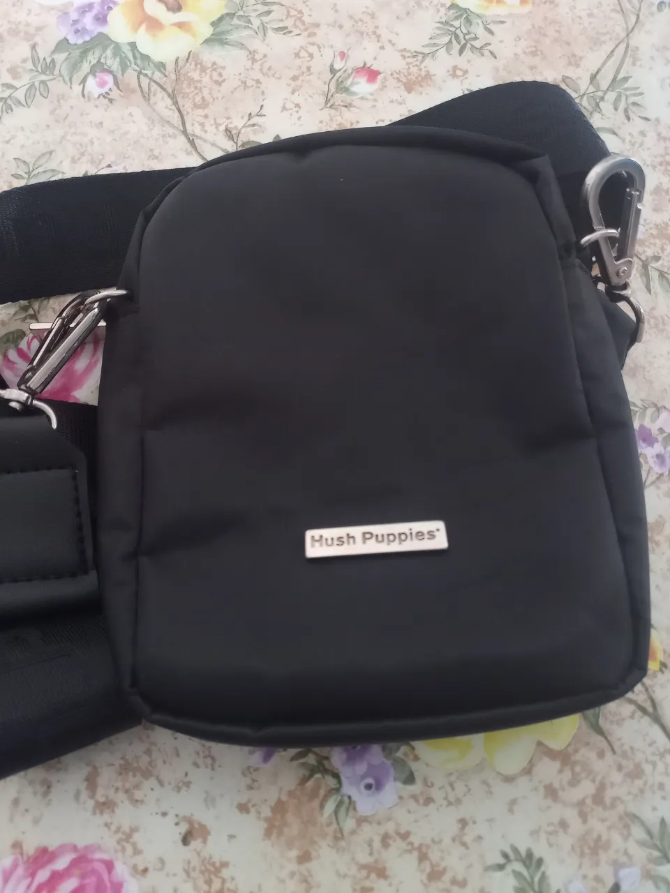 Bandolera negra Hush Puppies, ideal para llevar lo esencial. Confeccionada en material resistente, cuenta con un compartimento principal y un bolsillo frontal con cierre. Detalles de la marca en plateado. Perfecta para un look casual y moderno.