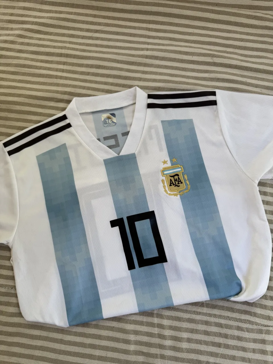 Remera de fútbol blanca con franjas verticales celestes y negras en los hombros. Estampado del número 10 en el frente y "MESSI 10" en la espalda. Escudo de la AFA bordado en el pecho.