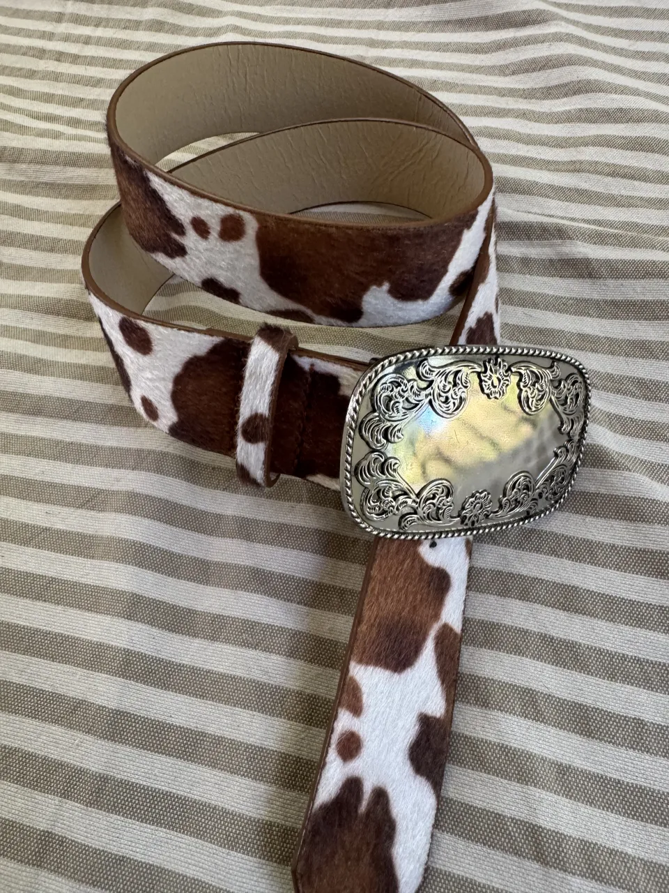 Cinturón con hebilla plateada y diseño grabado. La correa tiene un estampado de vaca en blanco y marrón.