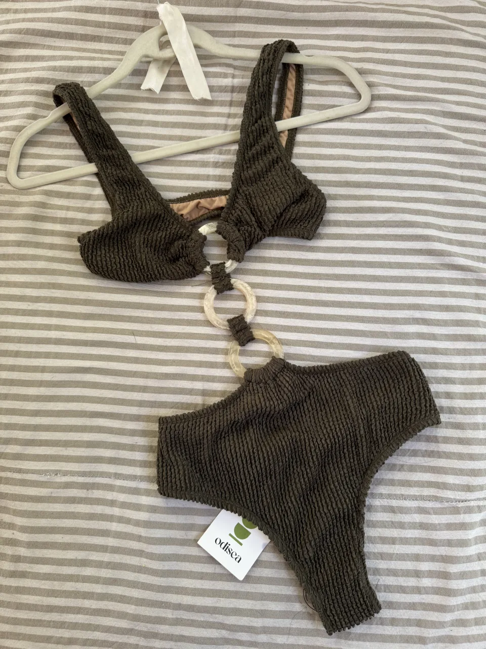 Traje de baño de dos piezas color verde oscuro. El corpiño es de estilo triangular con tiras finas y un detalle de argollas transparentes en el centro. La bombacha es tiro alto con detalles de argollas transparentes a los costados.