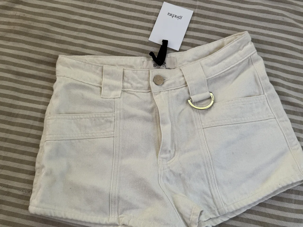 Shorts de gabardina color blanco tiza. Poseen presillas para cinturón y un detalle metálico en forma de D en la presilla izquierda. Tienen bolsillos frontales y traseros.