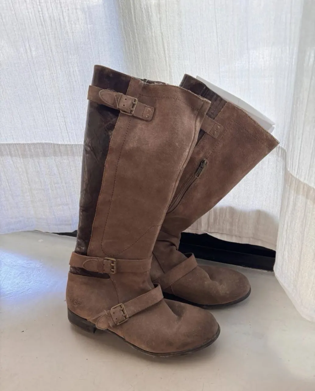 Botas marca UGG de caña alta de cuero gamuzado color marrón. Tienen hebillas decorativas y una tira con hebilla en la parte superior de la caña y otra en el tobillo. Cierre lateral.