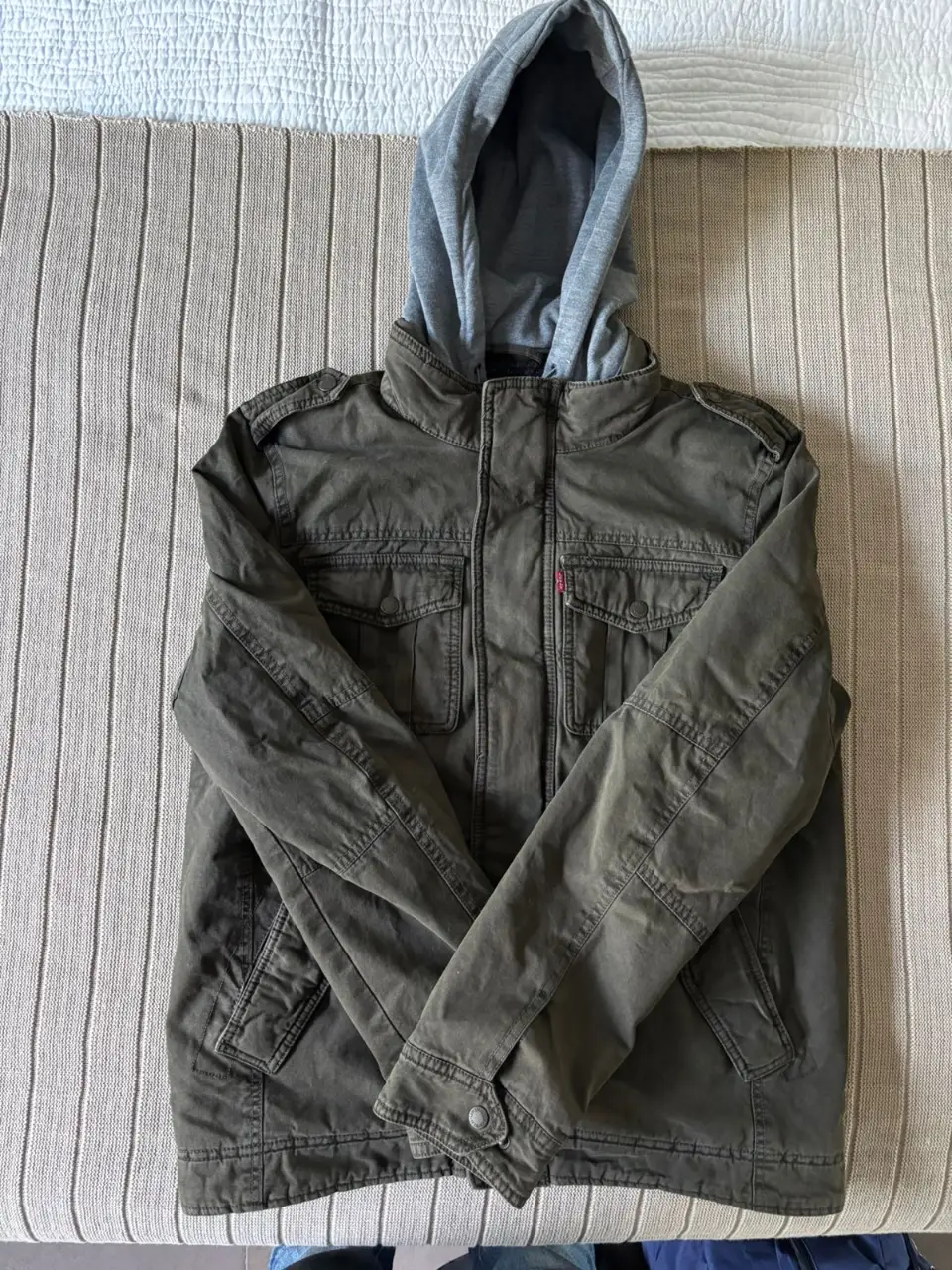 Campera militar verde con capucha gris desmontable. Posee bolsillos delanteros y cierre frontal. Ideal para un look casual y urbano.