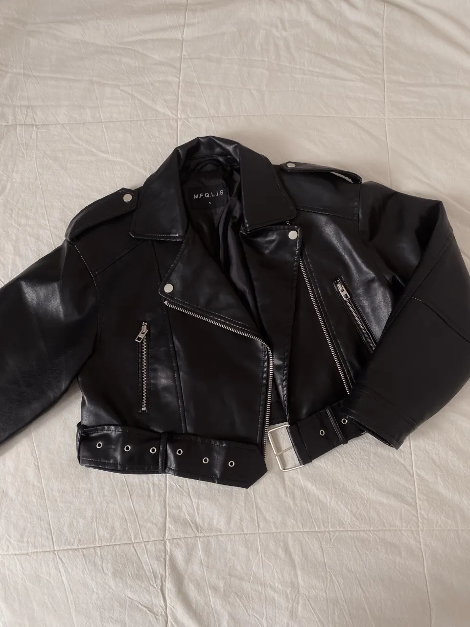 Campera de simil cuero  negra con cuello solapa, cierre metálico al frente, bolsillos con cierre y cinturón con hebilla metálica en la cintura.