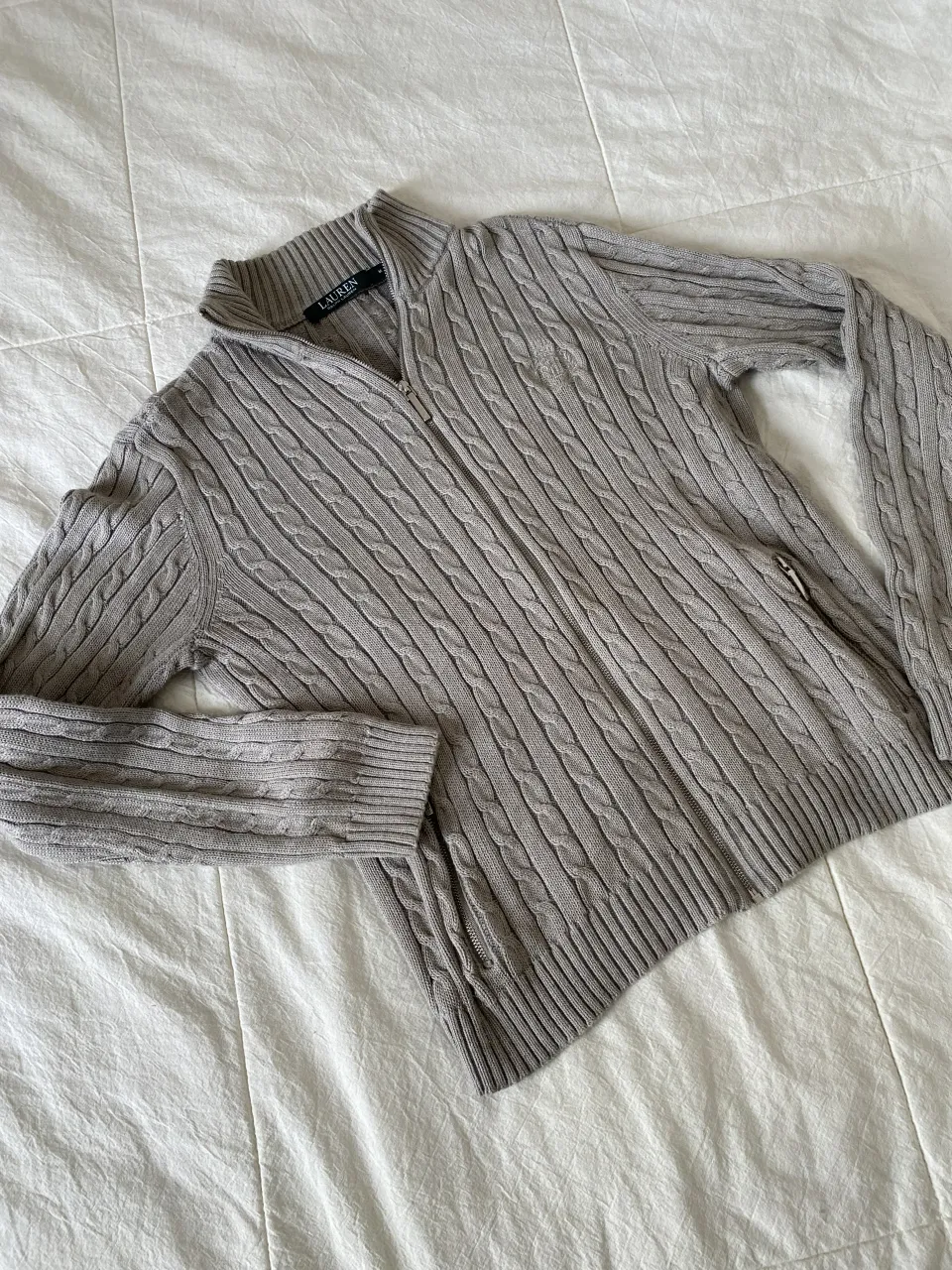 Sweater gris con cuello alto y cierre completo. Posee tejido de trenzas en toda la prenda. Igual a nuevo tiene un solo uso