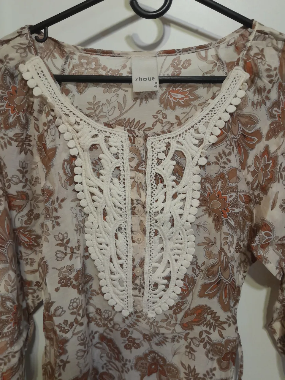 Blusa estampada con detalles de encaje y pompones en el cuello. Posee botones en la parte delantera y mangas largas.