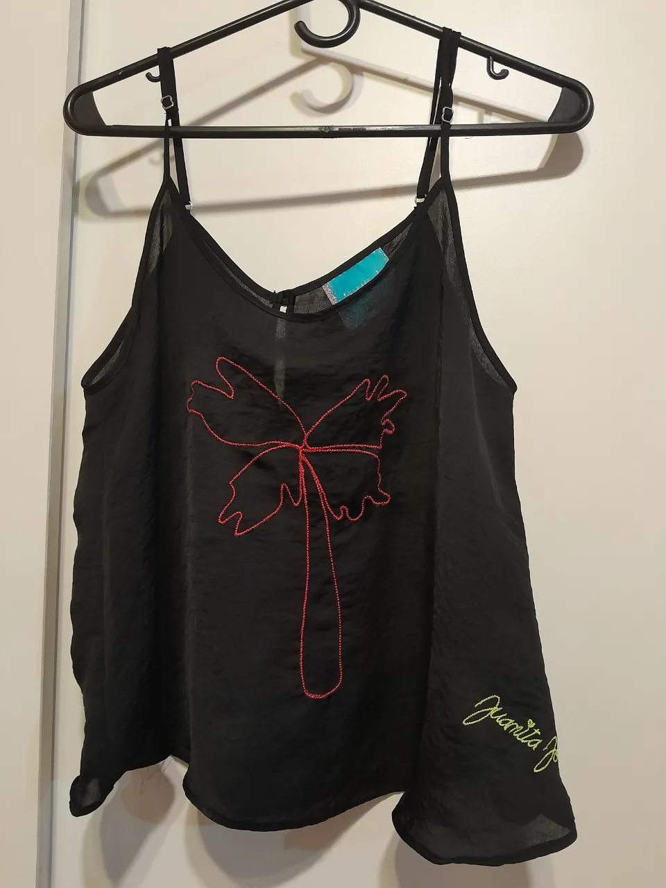 Musculosa negra con bordado de flor roja y firma "Juanita J" en amarillo. Ideal para un look casual y fresco.