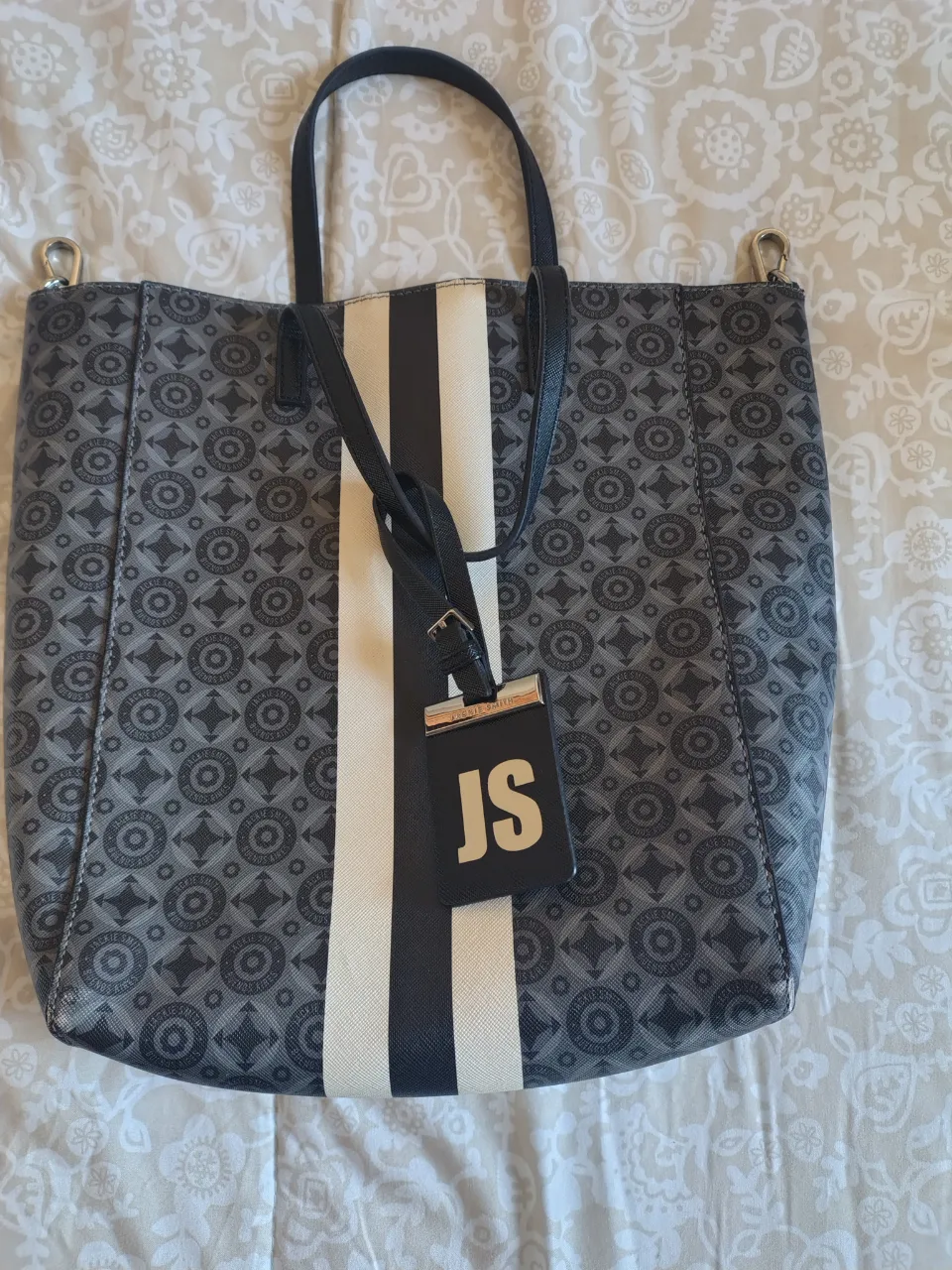 Bolso tote de mujer, con diseño geométrico en tonos grises y blanco. Cuenta con una franja central blanca y negra, manijas superiores y una correa ajustable para llevar al hombro. Incluye un llavero con las iniciales 'JS'. Ideal para uso diario.