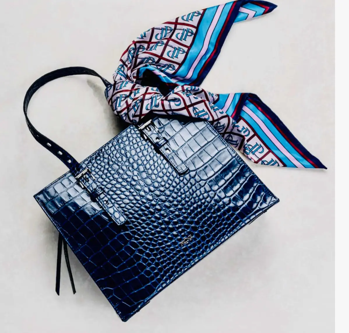Cartera de mano azul con textura de cocodrilo, ideal para un look elegante. Incluye correa para usar al hombro y un pañuelo estampado.