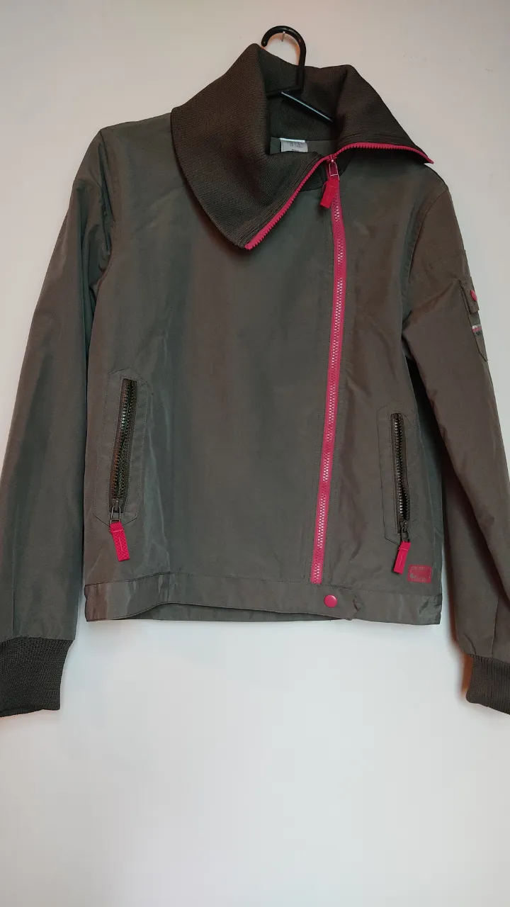 Campera de color caqui con cierre fucsia y cuello alto tejido. Posee bolsillos laterales con cierre y puños tejidos.
Pequeño bolsillo exterior con broches en una de las mangas. Interior forrado con microtoalla muy transpirable. 
Medidas: Contorno de busto 92cm. 
Espalda de hombro a Hombro 41cm. Largo total 56cm. Largo de manga 55cm