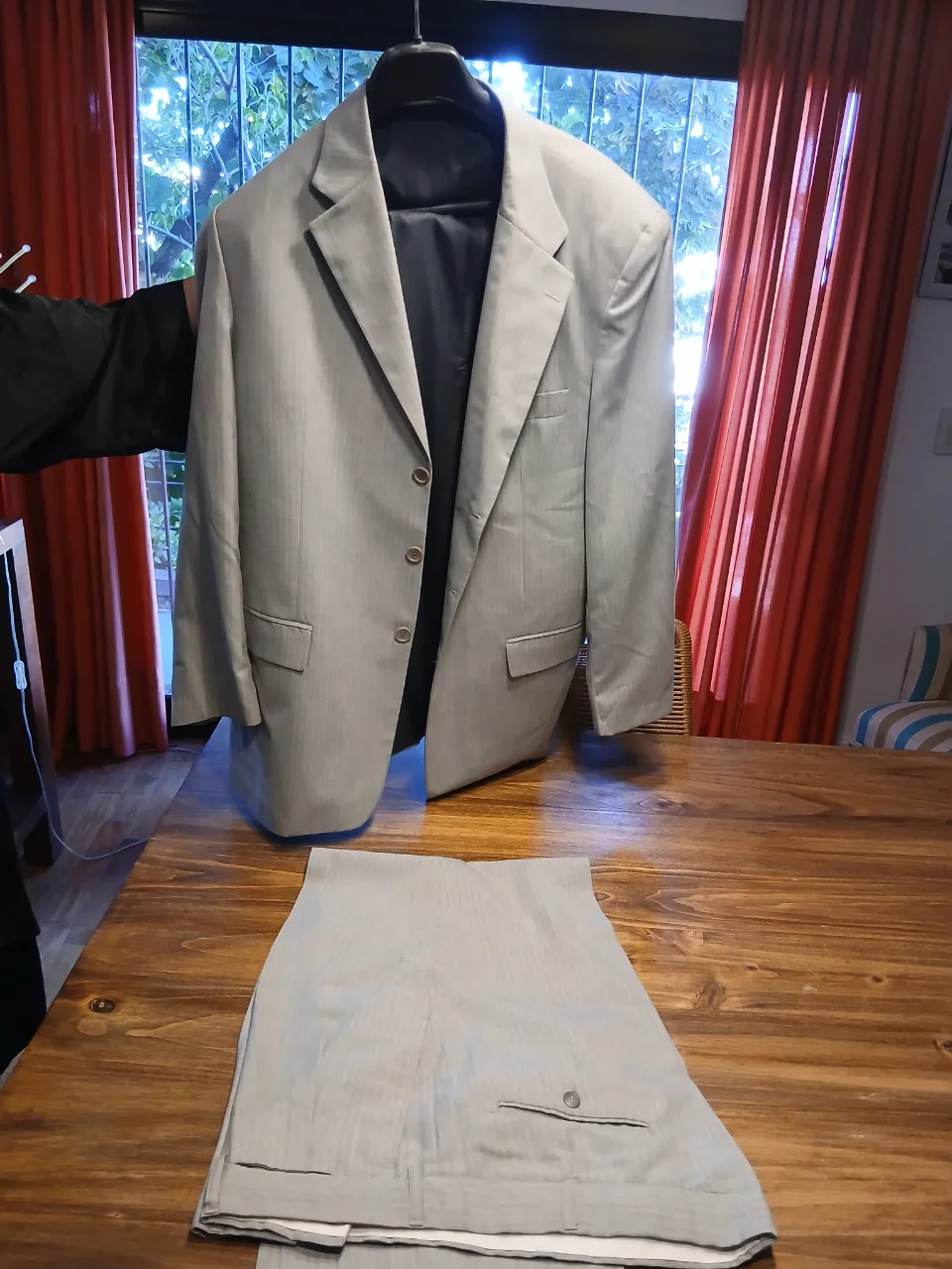 Traje de saco y pantalón gris claro con finas rayas verticales. El saco tiene solapas clásicas, tres botones al frente y bolsillos con tapa. El pantalón es liso con un botón y cierre.