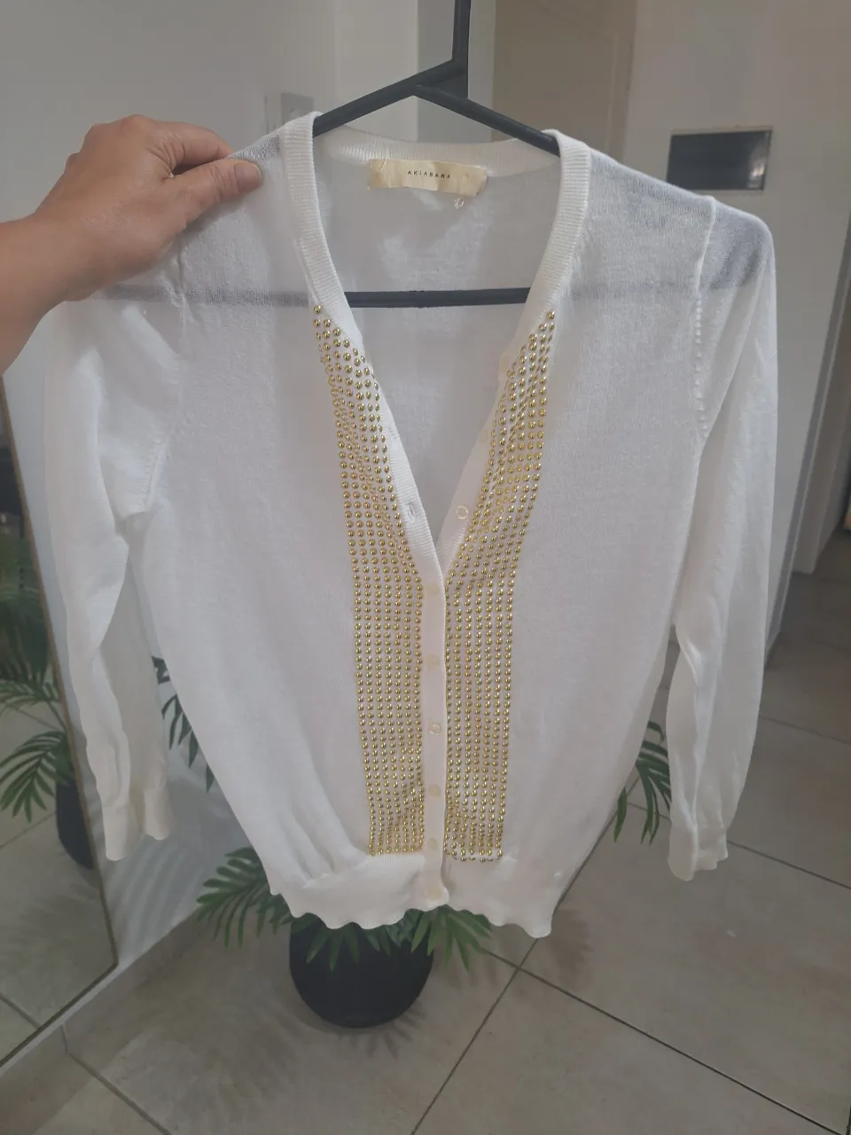 Cardigan blanco con detalles de tachas doradas en V y cuello. Manga larga y botones al frente. Un básico elegante para cualquier ocasión.