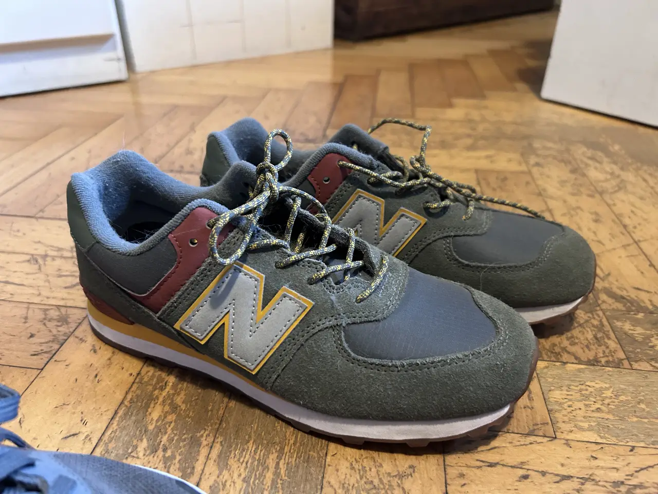 Zapatillas New Balance 574 para hombre, color verde y gris con detalles en amarillo y rojo. Ideales para uso diario y combinar con cualquier outfit casual.
