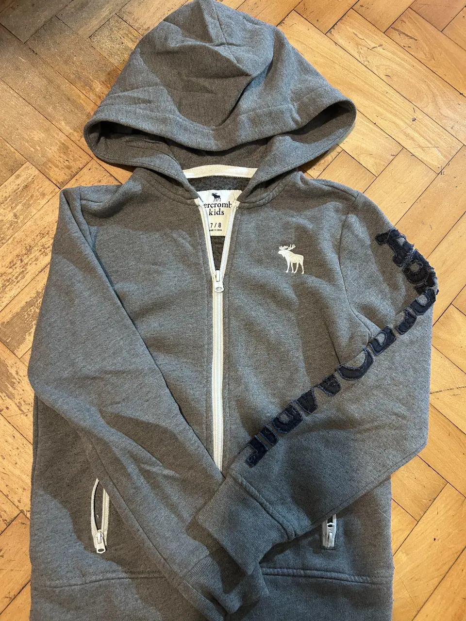 Buzo con cierre y capucha para niño, de la marca Abercrombie Kids. Talle 7/8. Con logo de la marca en el pecho y detalle de la marca en la manga.